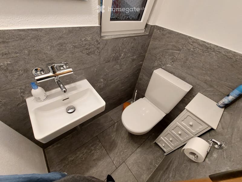 Modern bathroom with a white sink, chrome faucet, toilet, toilet paper holder, and a small stool. The walls are tiled in a grey marble pattern, and the floor is tiled in a similar grey marble design.