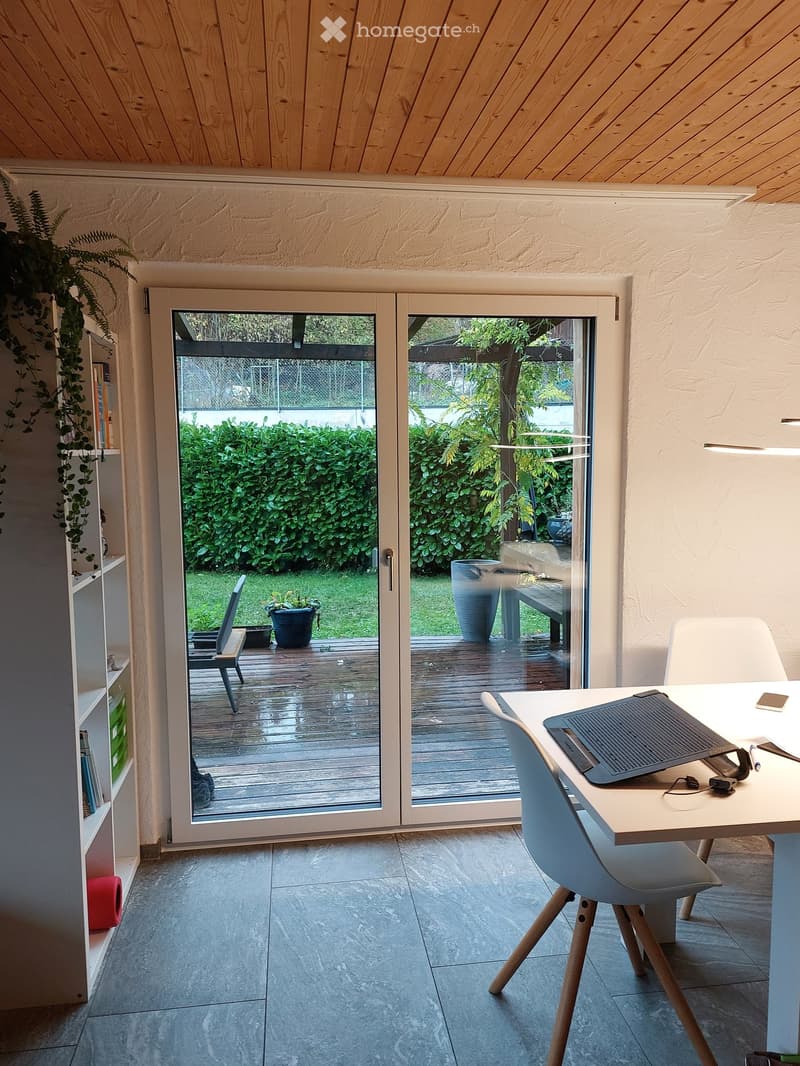 A modern, well-lit room with a desk, chair, laptop, and a large window opening to a wooden deck and garden. The room features a tiled floor, a wooden ceiling, and a shelf with books and plants.