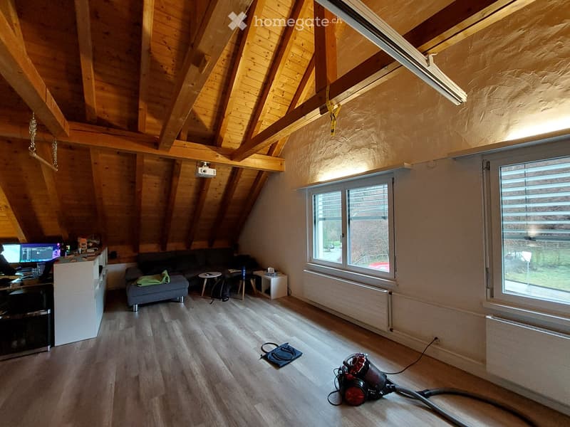 Loft-style living room with wooden beams, exposed ceiling, large windows, sofa, small table, laptop, and vacuum cleaner on the floor.