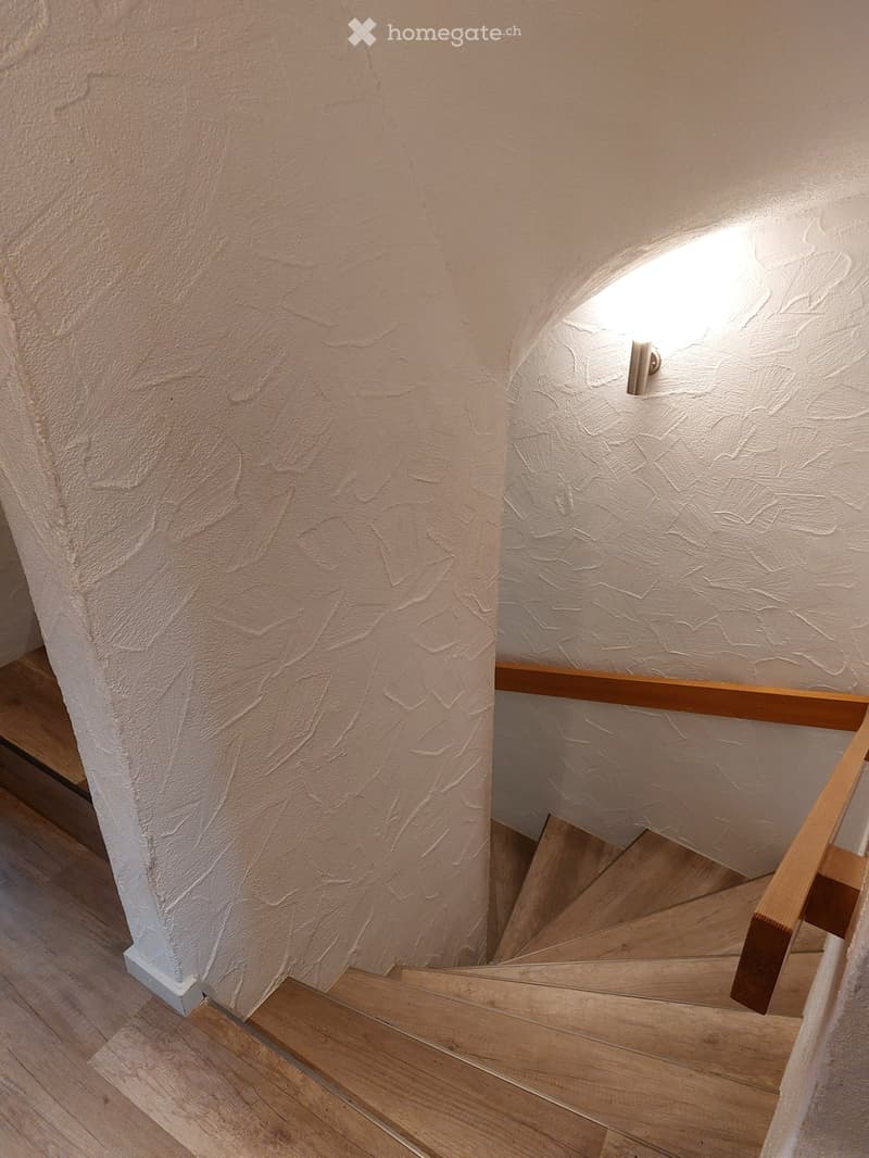 Wooden staircase with a wooden handrail and white textured walls, illuminated by a small light fixture on the wall.