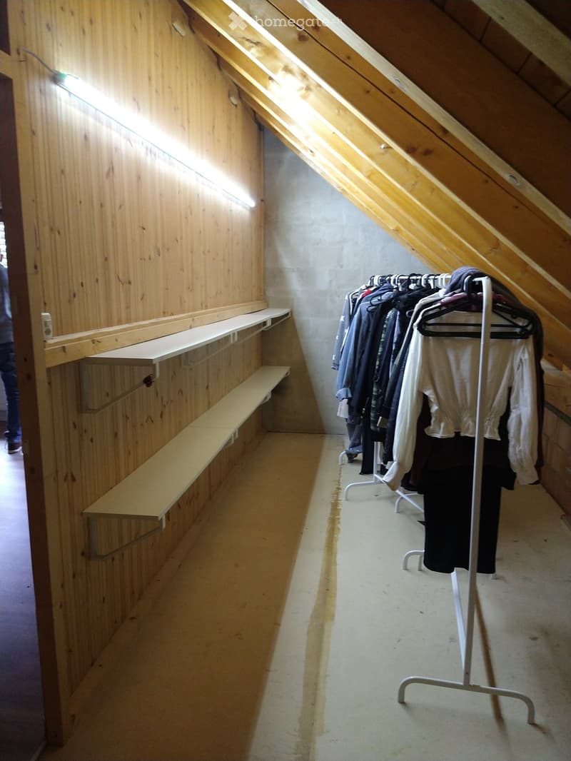 A narrow hallway with wooden walls and built-in wooden shelves on one side, and a clothing rack with hanging garments on the other side.