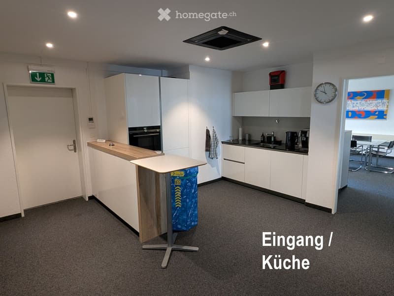 Modern kitchen with white cabinets, a wooden counter, sink, oven, and a small table with chairs. A clock is mounted on the wall, and a trash bin is visible.