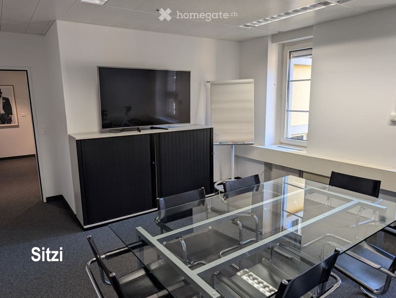 Modern office or meeting room with a large glass table surrounded by black chairs, a mounted screen on the wall, and a cabinet with a framed picture on the side.