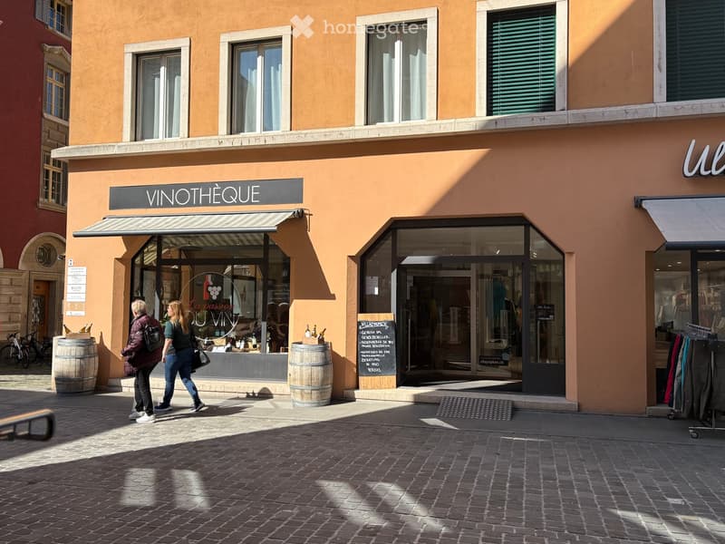 A two-story building with a wine shop on the ground floor, featuring a glass door, a blackboard with text, and wooden barrels outside. The building has several windows and a sign reading 'VINOTHEQUE' above the entrance.