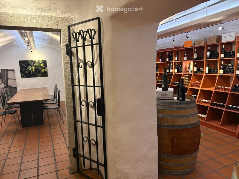 A room with a wooden table, chairs, a black gate, a barrel, shelves with wine bottles, and a painting on the wall.