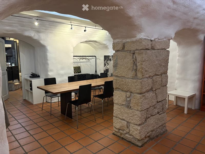A room with a long wooden table, black chairs, a white shelf, a stone pillar, and a tiled floor.