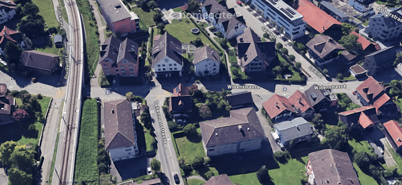 Aerial view of a residential area with multiple houses, streets, and a railway line. Houses have varied roof styles and colors, surrounded by greenery and trees.