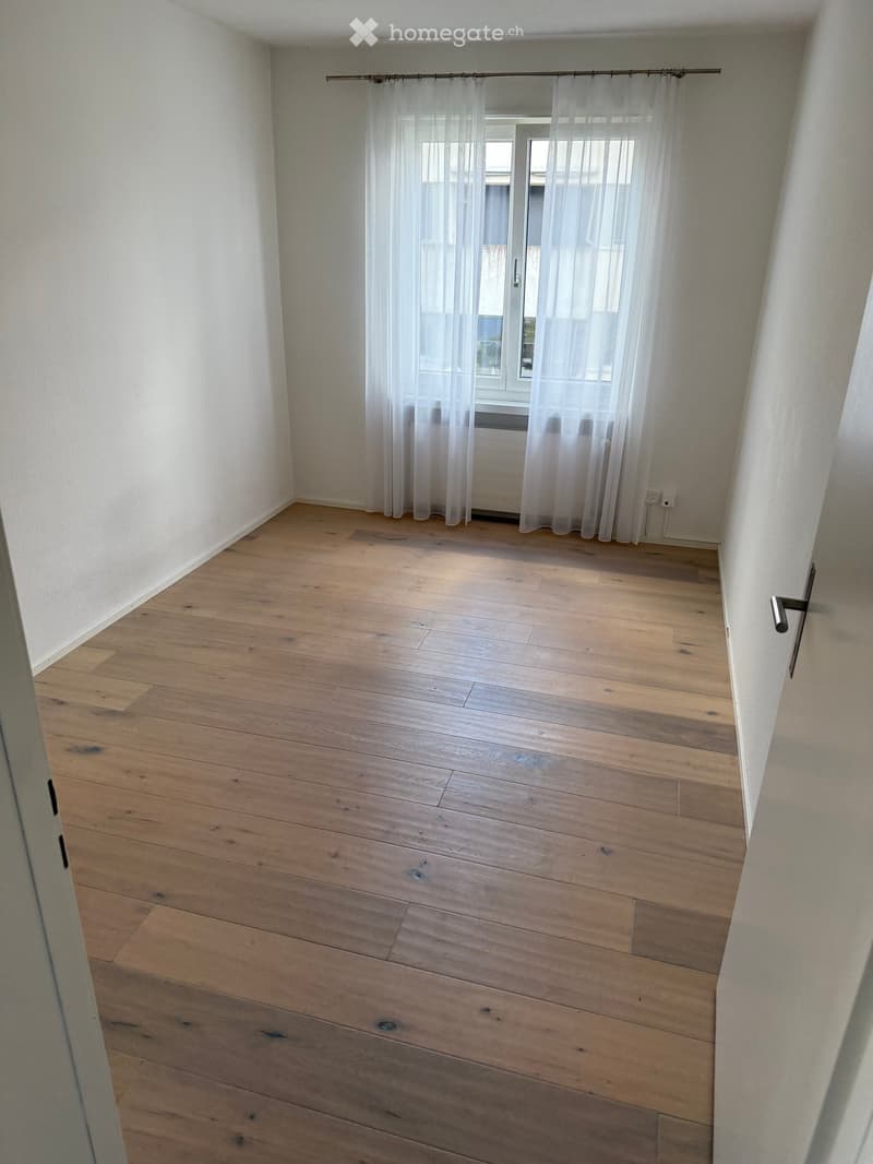 Empty room with light wooden flooring, white walls, and a window with sheer curtains.