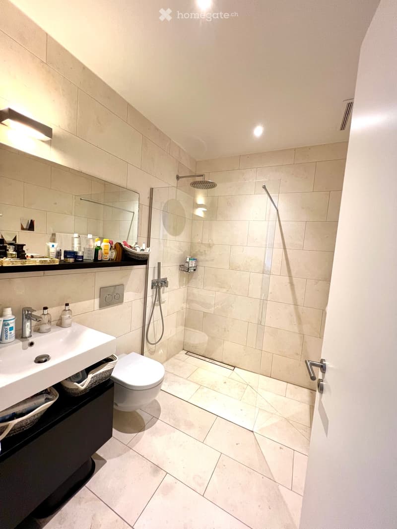 Modern bathroom with a white sink, toilet, and glass-enclosed shower. The room features beige tiles, a large mirror, and various toiletries on the sink counter.