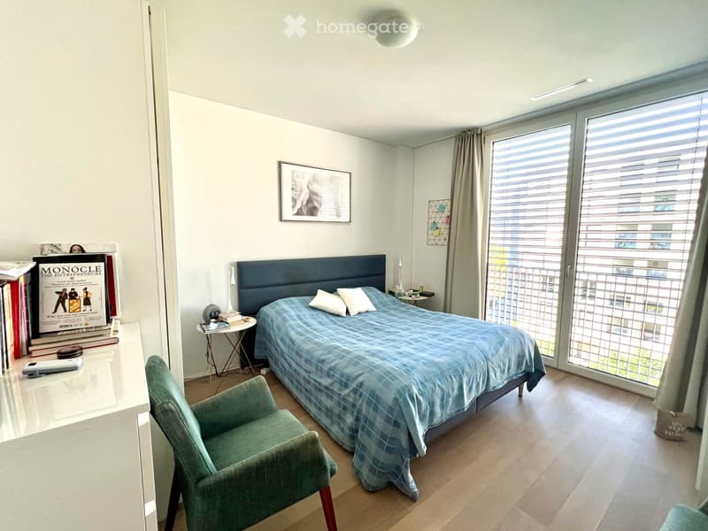 Modern bedroom with a blue checkered bedspread, a green armchair, a nightstand, a small table with books, and a large window with blinds.