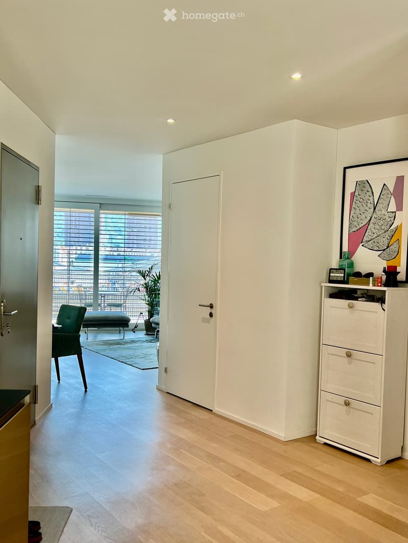 Modern hallway with wooden flooring, white walls, and a view of a cityscape through large windows. Features a green chair, a small stool, a white cabinet, and a decorative picture frame.