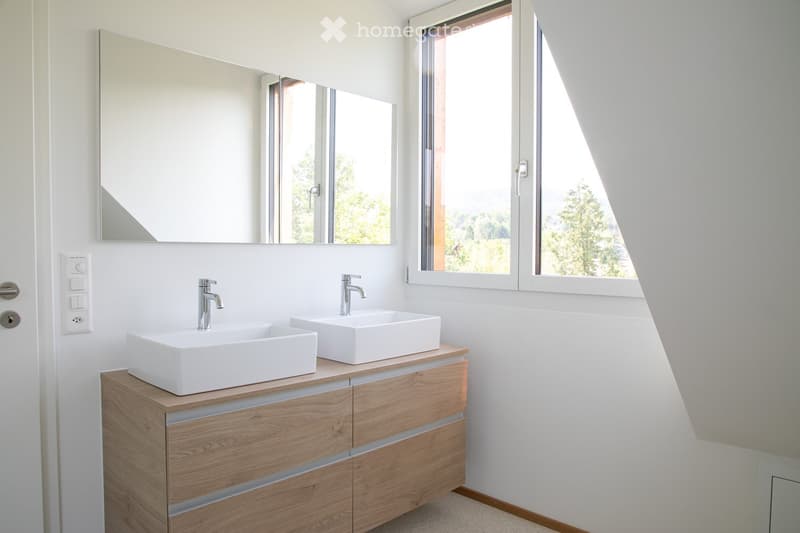Modern bathroom with two white sinks on a wooden vanity, large mirror, and large window offering a view of greenery.