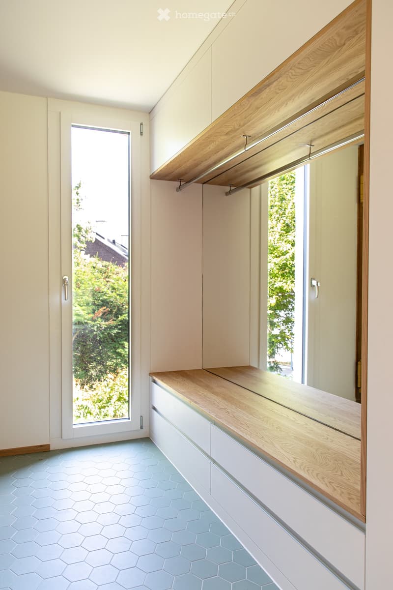 Modern hallway with light-colored walls, hexagonal light blue tiles, built-in wooden bench seating, and large windows offering a view of greenery outside.