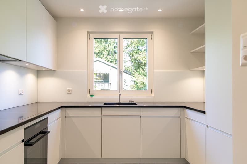 Modern kitchen with white cabinets, black countertops, a sink, and an oven. Large windows provide natural light.
