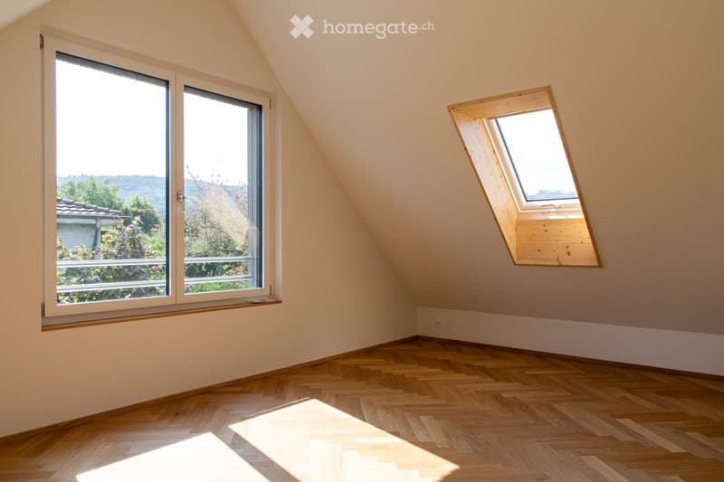 Empty attic room with wooden flooring, slanted ceilings, large windows, and a skylight. No furniture or equipment present.