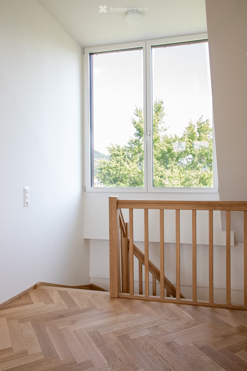 Empty hallway with wooden flooring, white walls, and a wooden railing leading to a window with a view of greenery outside.
