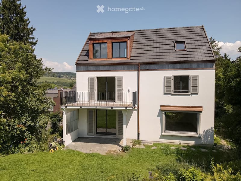 Two-story modern house with white exterior, grey roof, balcony, large windows, and lush green surroundings.