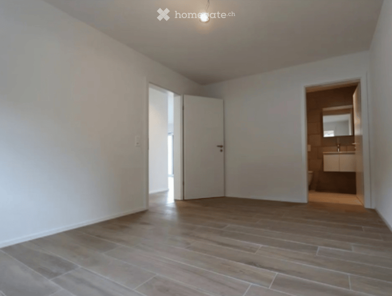 Empty room with wooden flooring and white walls, featuring an open doorway leading to a bathroom.