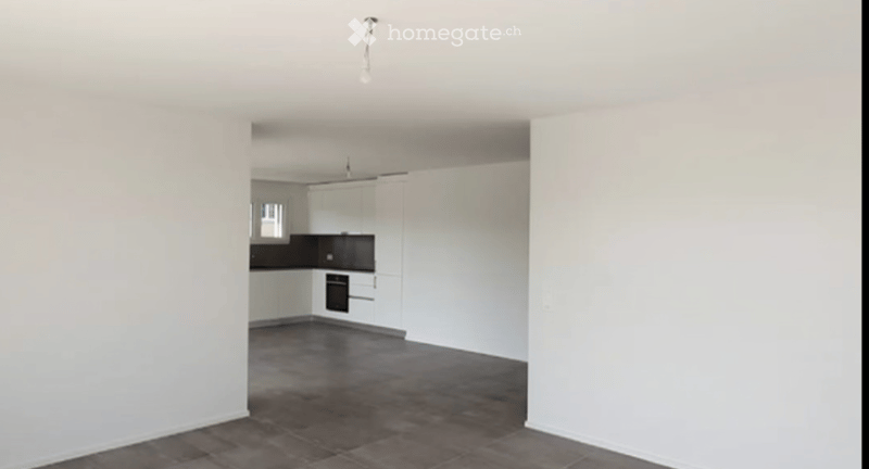 Empty room with white walls, dark tiled floor, and an adjacent kitchen area with white cabinets and black countertops.