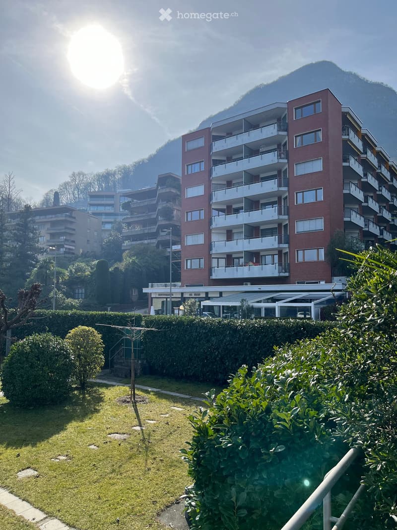 Multi-story apartment building with balconies, surrounded by lush greenery and gardens. The building has a modern, brick facade and is situated in a scenic location with mountains in the background.