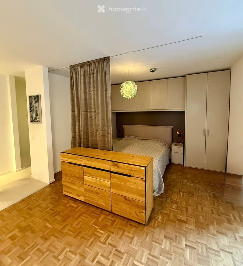 Modern bedroom with wooden flooring, a bed with a headboard, a nightstand, a dresser, built-in cabinets, and a curtained window.