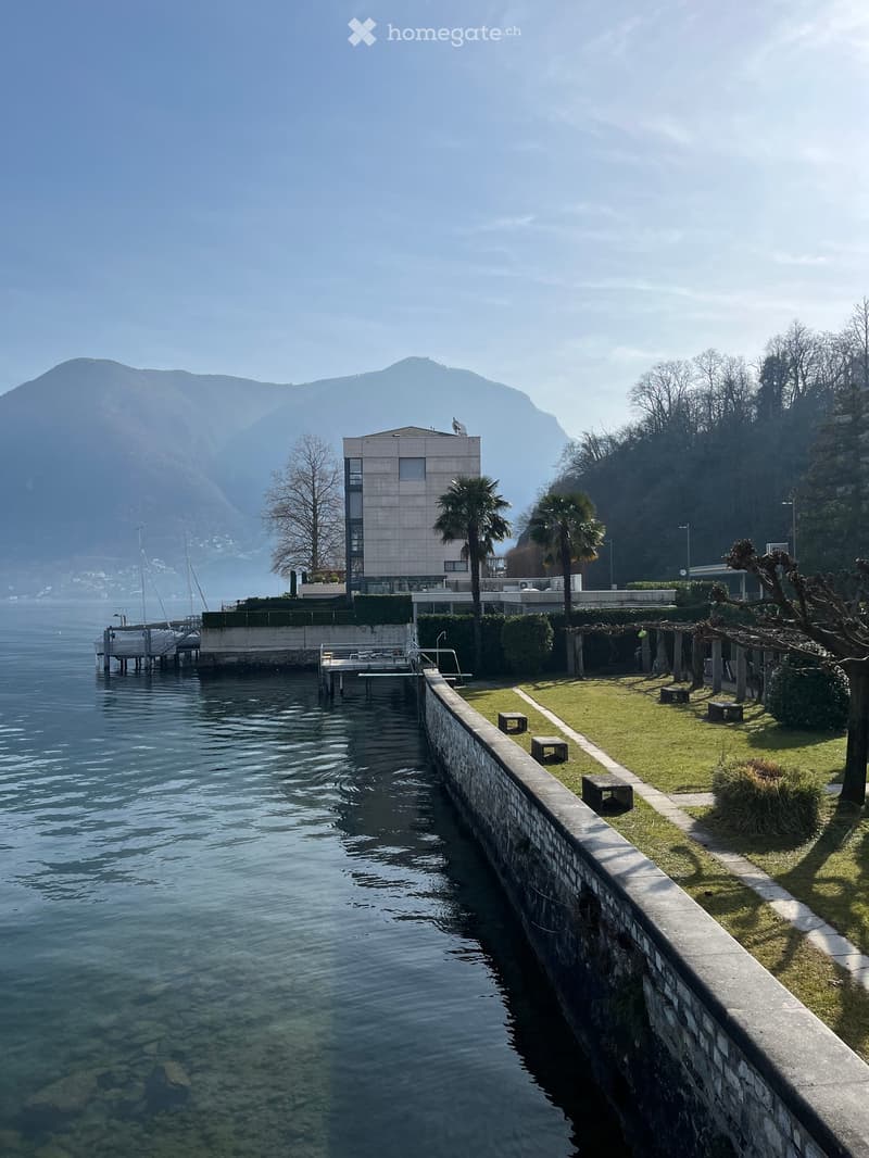 The image shows a lakefront property with a modern building, a dock, and a grassy area along the water's edge. The building appears to be a hotel or apartment complex, with palm trees and other landscaping around it. The lake is surrounded by mountains in