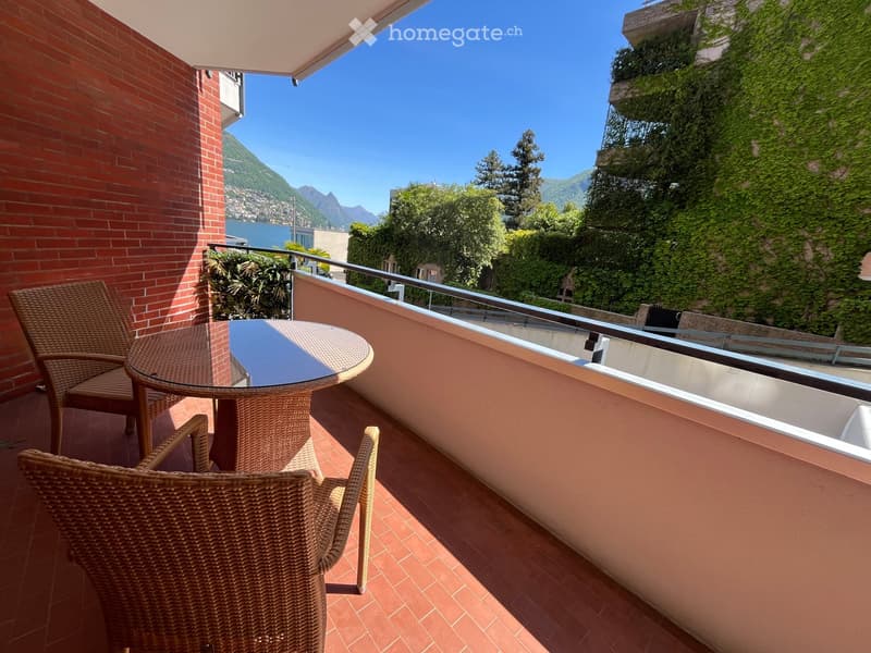 Balcony with a round glass table and wicker chairs, overlooking a scenic lake and mountain view.