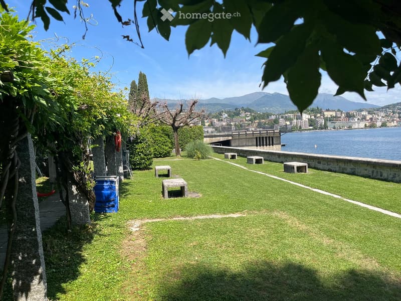 Well-maintained lawn with scattered stone benches, trees, and a scenic view of a lake and distant mountains.