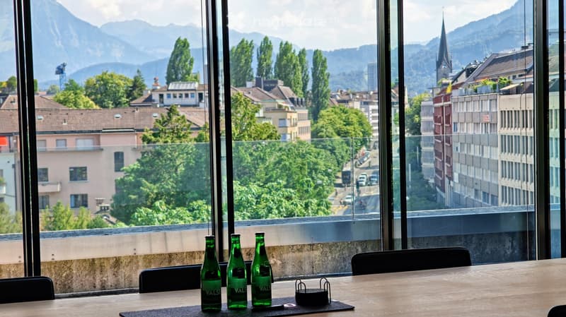 A modern room with large windows offering a scenic view of a cityscape and mountains, featuring a wooden table with three green bottles and several black chairs.