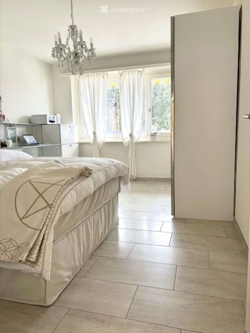 Bright bedroom with a large bed, chandelier, large windows with curtains, nightstand, and a mirror. The room features tiled flooring and a modern aesthetic.