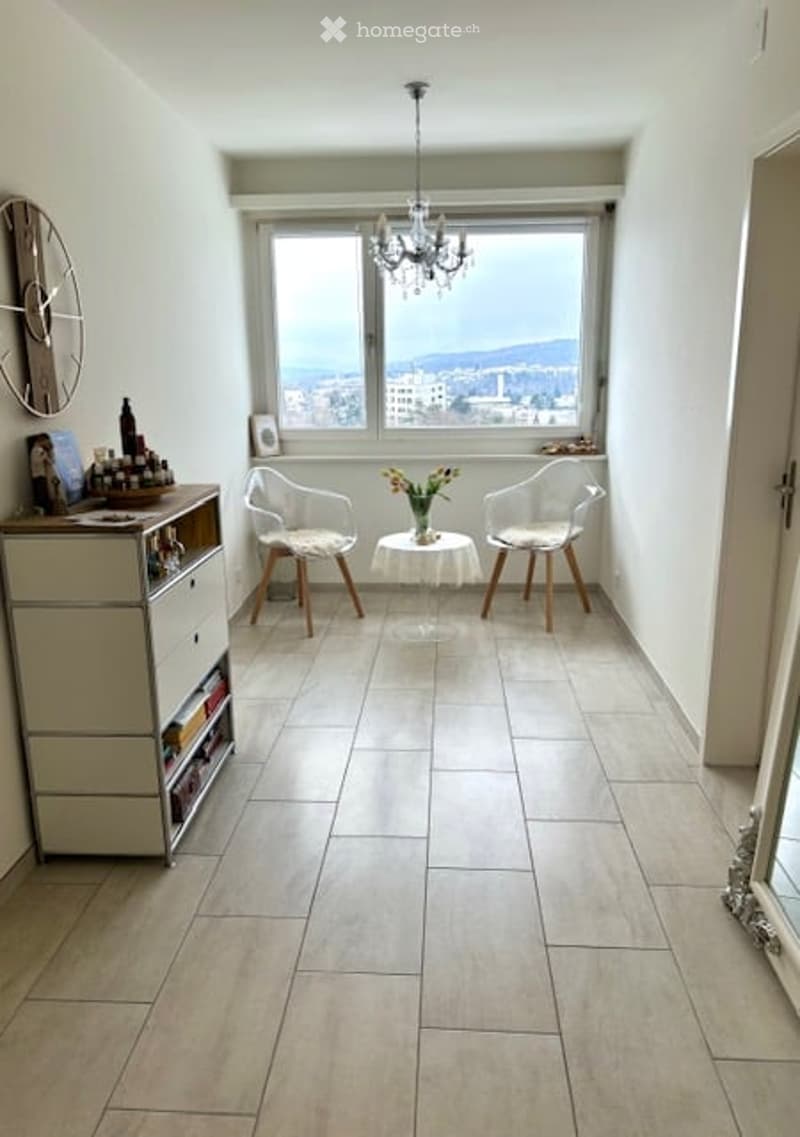 Bright living room with large window offering a city view, featuring two white chairs, a small round table, a cabinet with decorative items, and a chandelier.