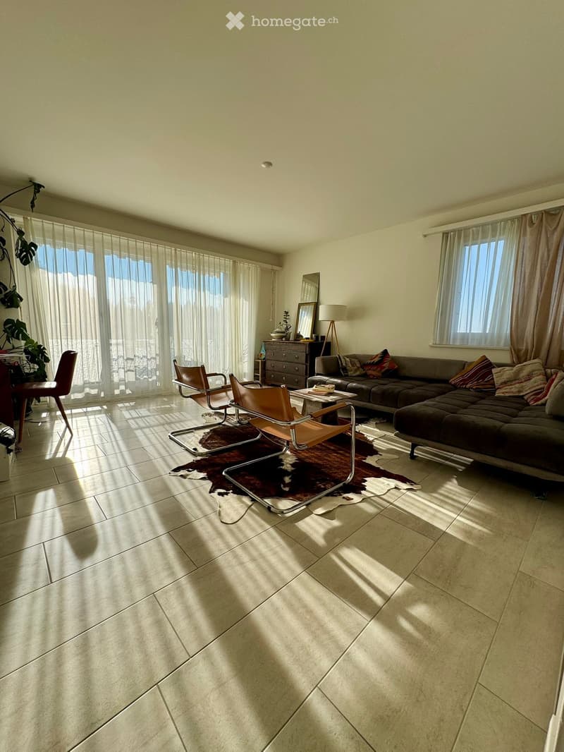 Bright living room with large windows, beige walls, tiled floor, and modern furniture including a sofa, chairs, a coffee table, and a lamp.
