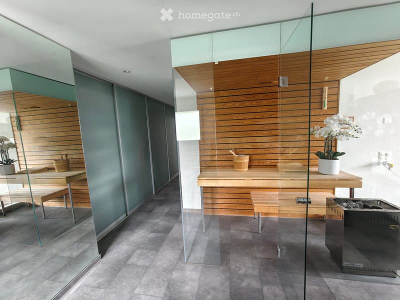 Modern bathroom with wooden sauna benches, a wooden bucket, a glass partition, and a flower pot on a table.