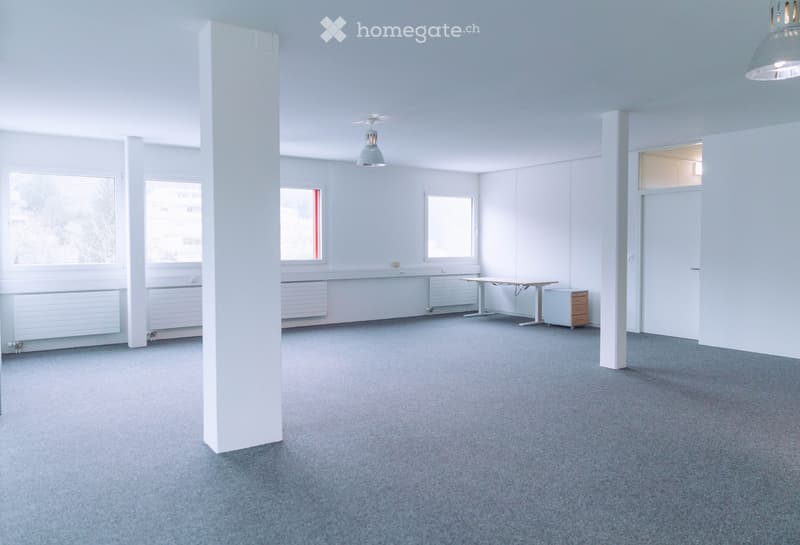 Bright, open room with grey carpeting, white walls, large windows, and a central table with a small cabinet nearby.