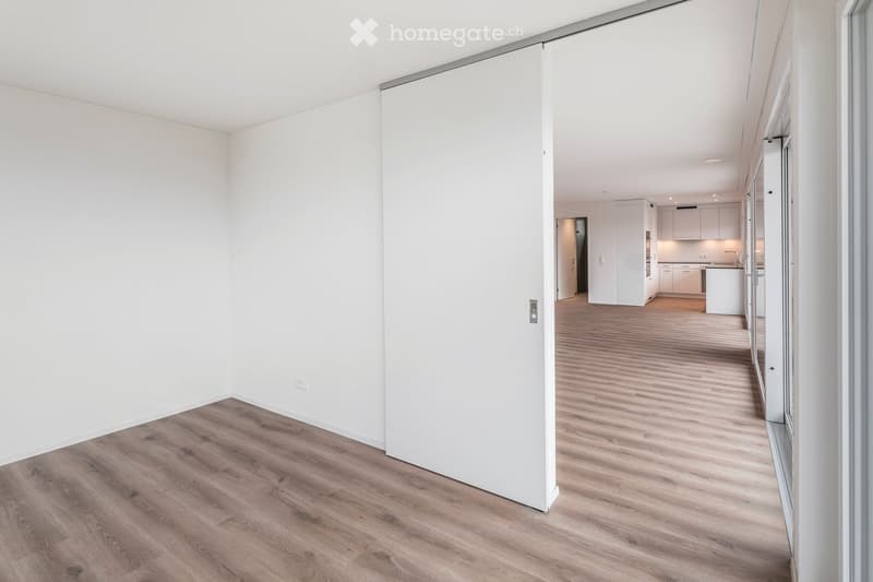 Empty room with white walls and wooden flooring, featuring a sliding door leading to another room with white cabinets and a kitchen area.