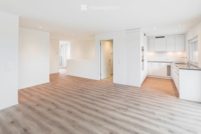 Empty room with light wooden flooring, white walls, and a modern kitchen area with white cabinets and stainless steel appliances visible in the background.