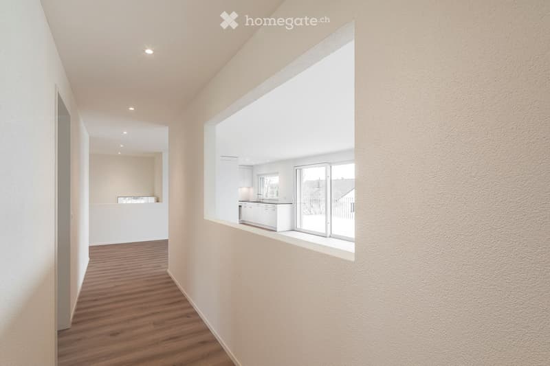 Bright, modern hallway with wooden flooring, recessed lighting, and a large window overlooking a kitchen area with white cabinets and a sliding glass door leading outside.