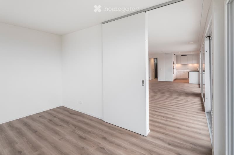 Empty room with white walls and wooden flooring, featuring a sliding door leading to a kitchen area with white cabinets and countertops.