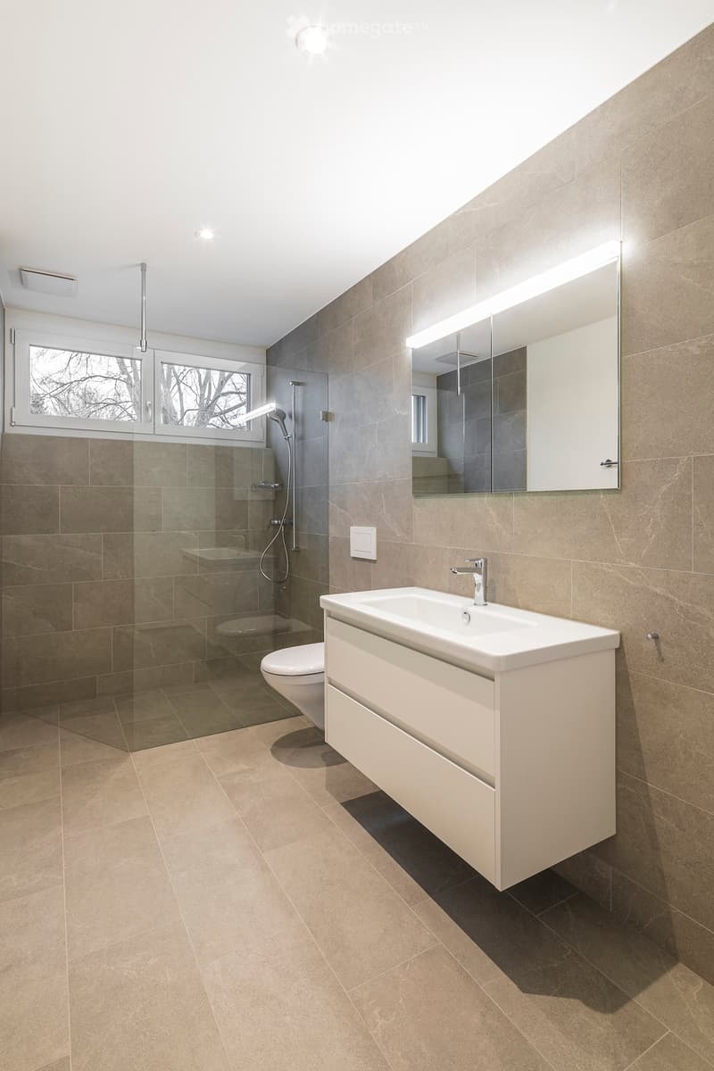 Modern bathroom with beige tiles, a white sink with a modern faucet, a large mirror with built-in lighting, a glass-enclosed shower, and a toilet.