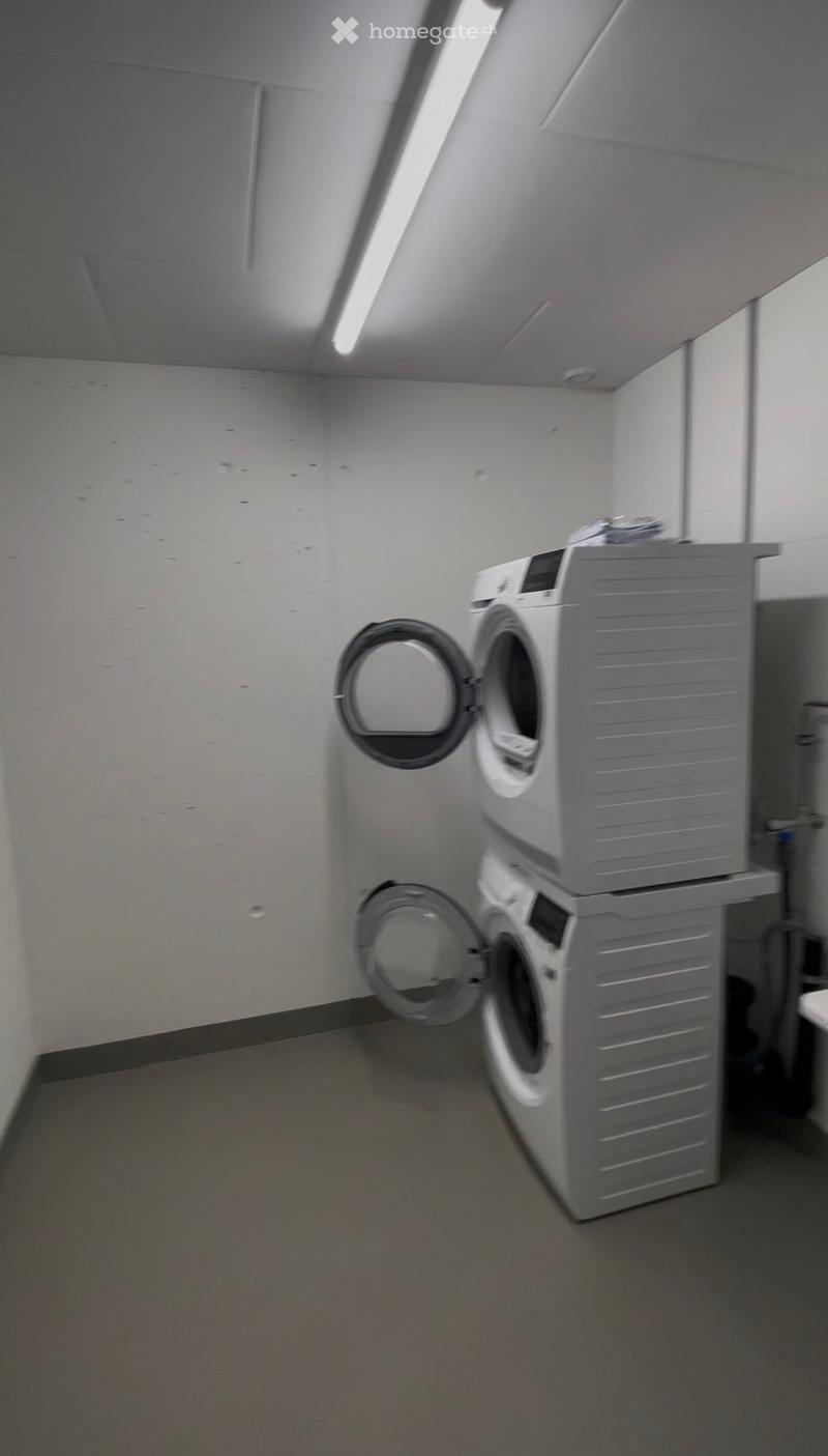 A small, well-lit room with a washing machine and dryer stacked together, white walls, and a grey floor.