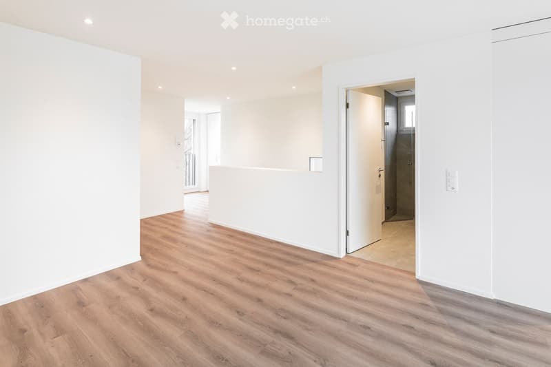 Empty room with light wooden flooring and white walls, featuring an open doorway leading to another room with tiled flooring.