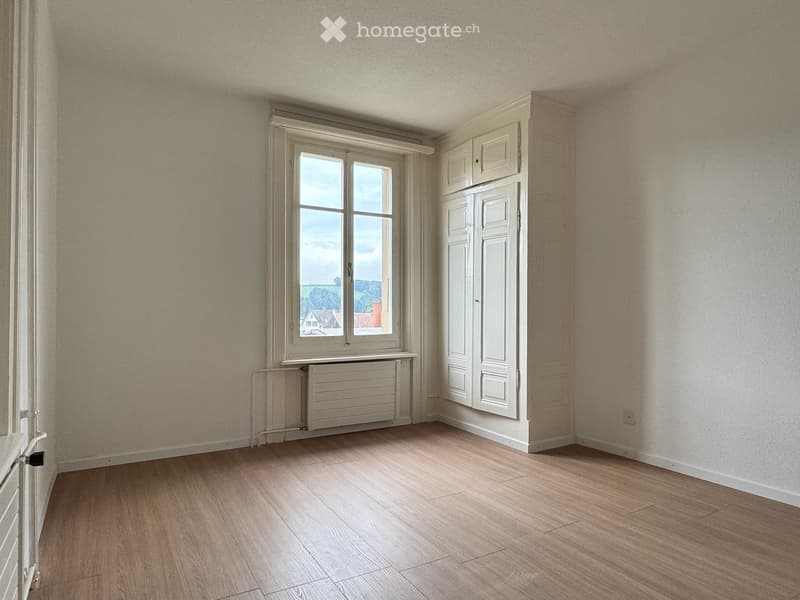 Empty room with wooden floors, white walls, large window with white frames, radiator, white wardrobe