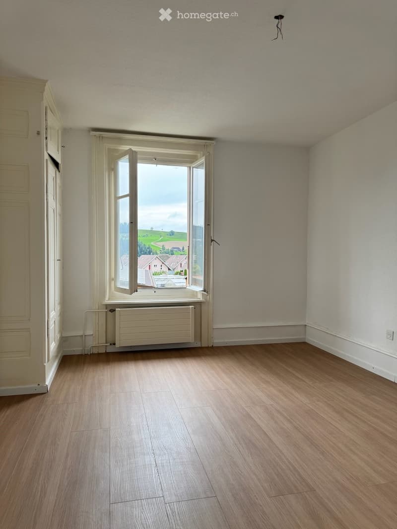 an empty room with white walls, wooden floor, and an open window showing a view of houses and greenery outside