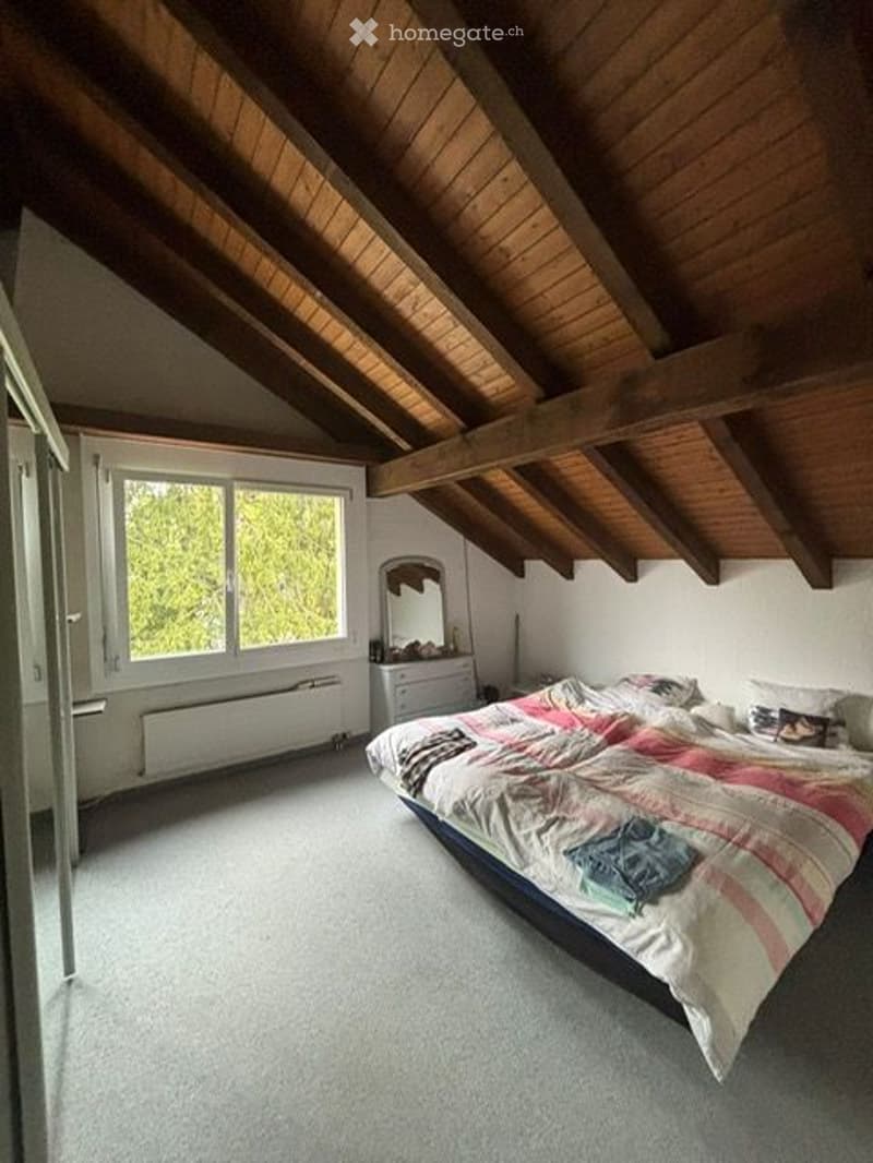 Cozy attic bedroom with wooden slanted ceilings, large window, bed with colorful bedding, dresser, and mirror.