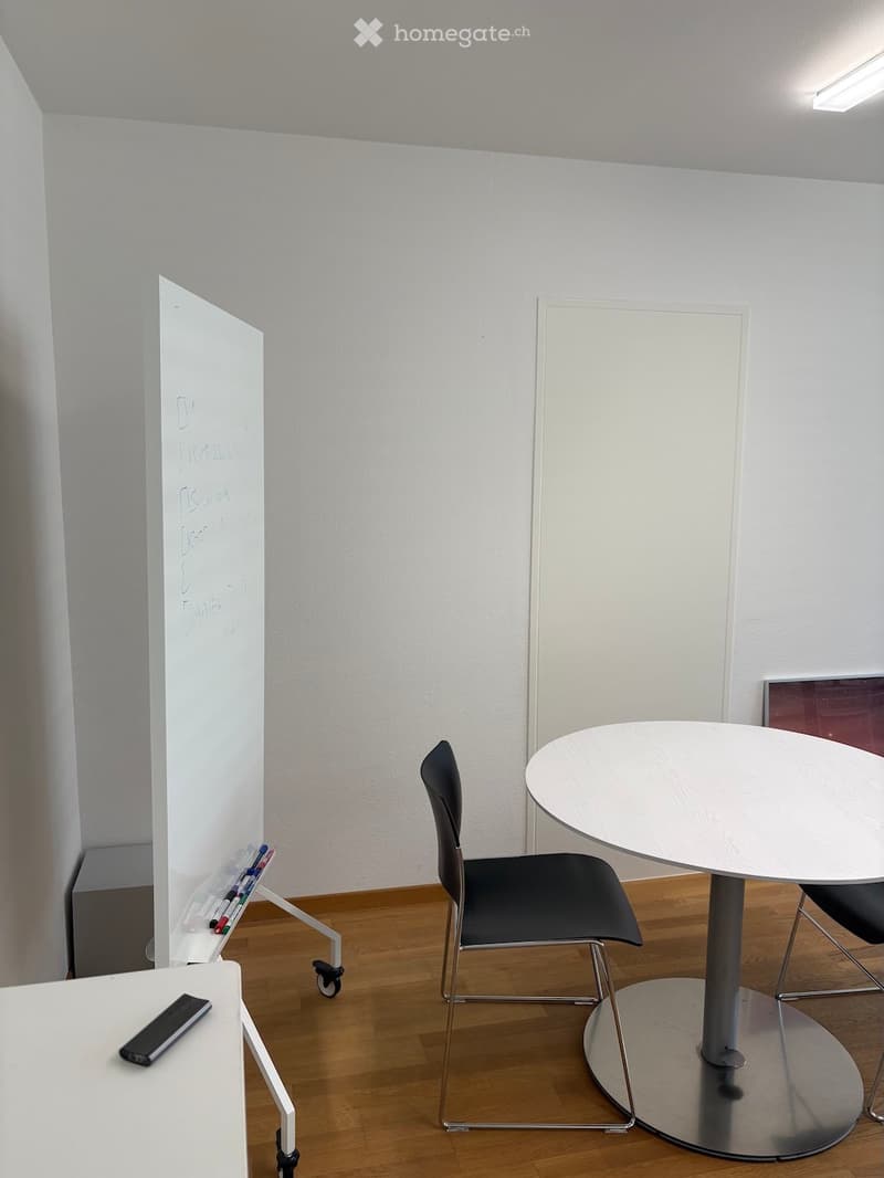 A modern, minimalist room with a round white table, black chair, whiteboard, and a small screen on a stand. The floor is wooden, and the walls are white.