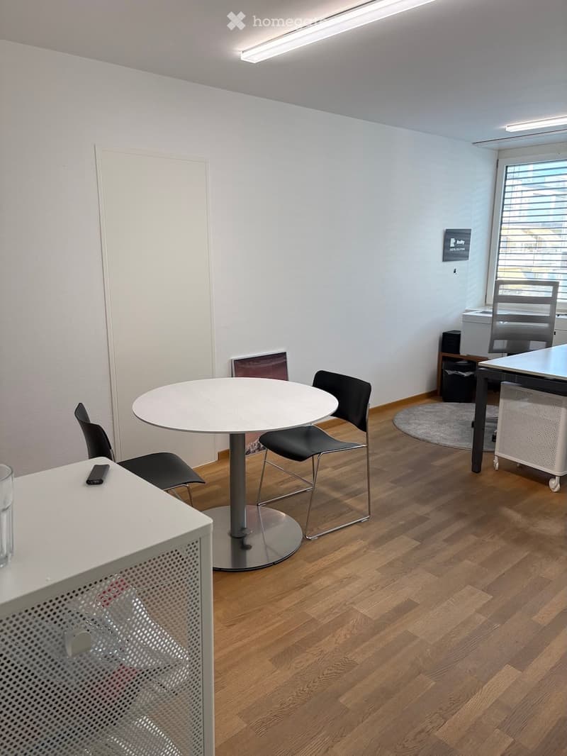 A modern, well-lit room with wooden flooring, a round white table with four black chairs, a white desk with a chair, and a small cabinet with a screen on top.