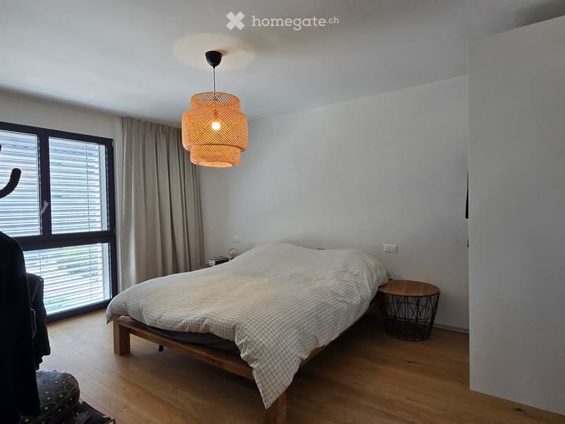Modern bedroom with a wooden bed, nightstand, small round table, and a hanging woven lamp. Large window with blinds and curtains allows natural light to enter.