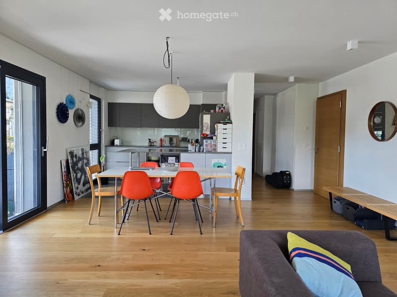 Open-plan living and dining area with wooden flooring, a grey sofa with colorful pillows, a white dining table with orange chairs, a modern kitchen with black cabinets and white countertops, and a hanging spherical lamp.