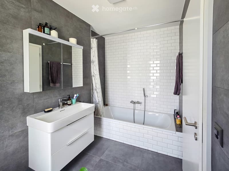 Modern bathroom with white tiles, a white sink, a bathtub, a large mirror, and various toiletries.