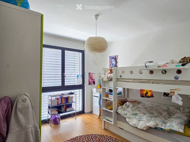 A modern, well-lit bedroom with a bunk bed, bookshelf, and decorative items. The room features a large window with blinds, a colorful rug, and a cozy atmosphere.